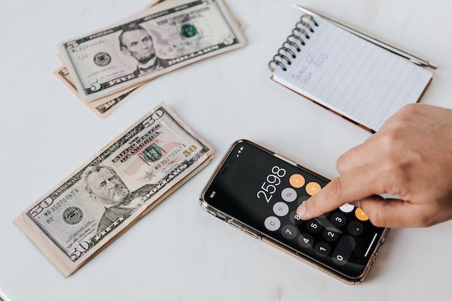 investing-37 Crop unrecognizable financier using calculator on smartphone near dollar banknotes